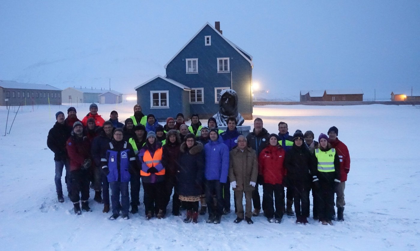 NySMAC members gathered in the snow in front of a blue wooden house in Ny-Ålesund